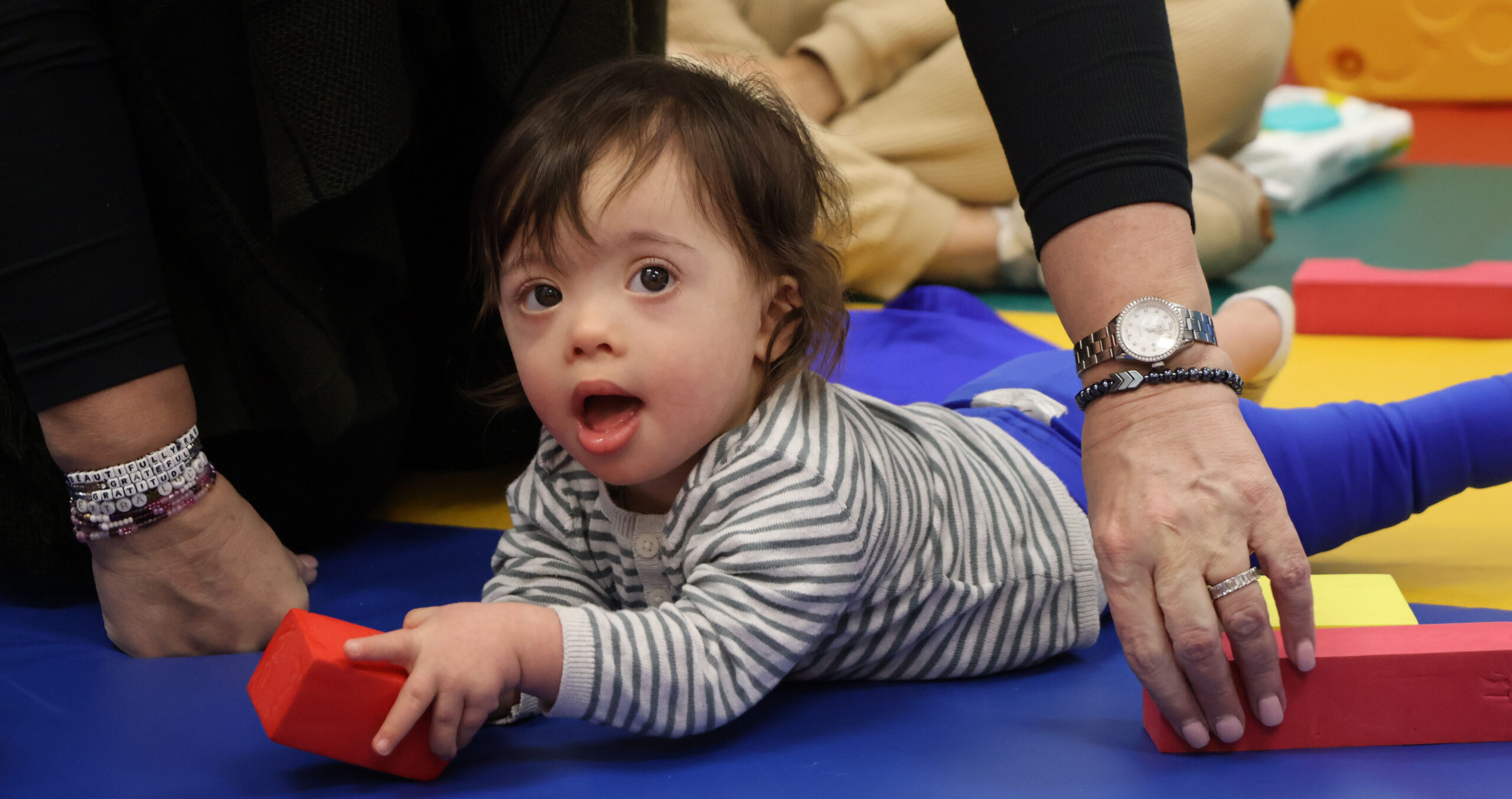 A baby boy is on his stomach on a soft floor mat, holding a toy.