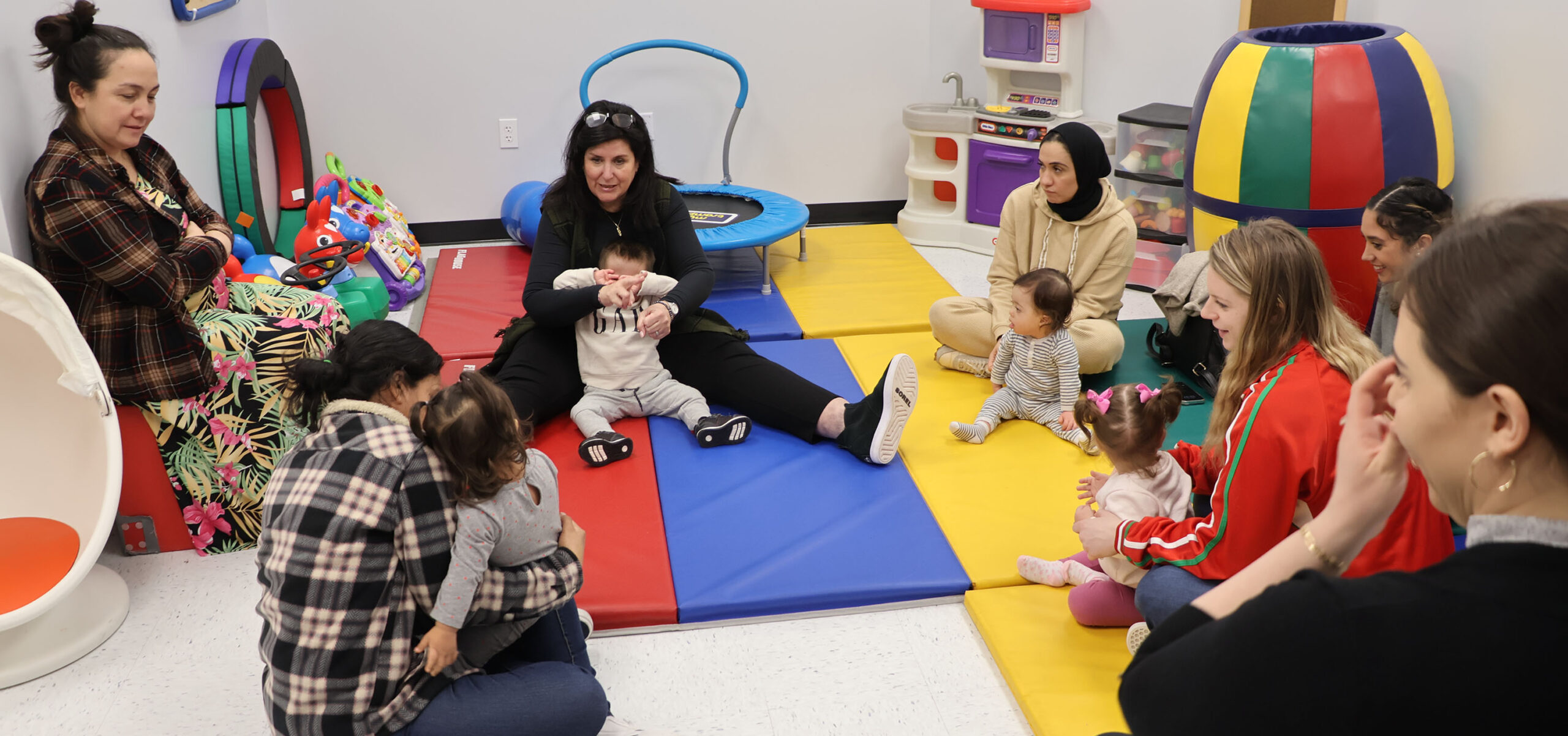 A group of women sit in a circle with young babies and toddlers, and playful equipment is all around them.