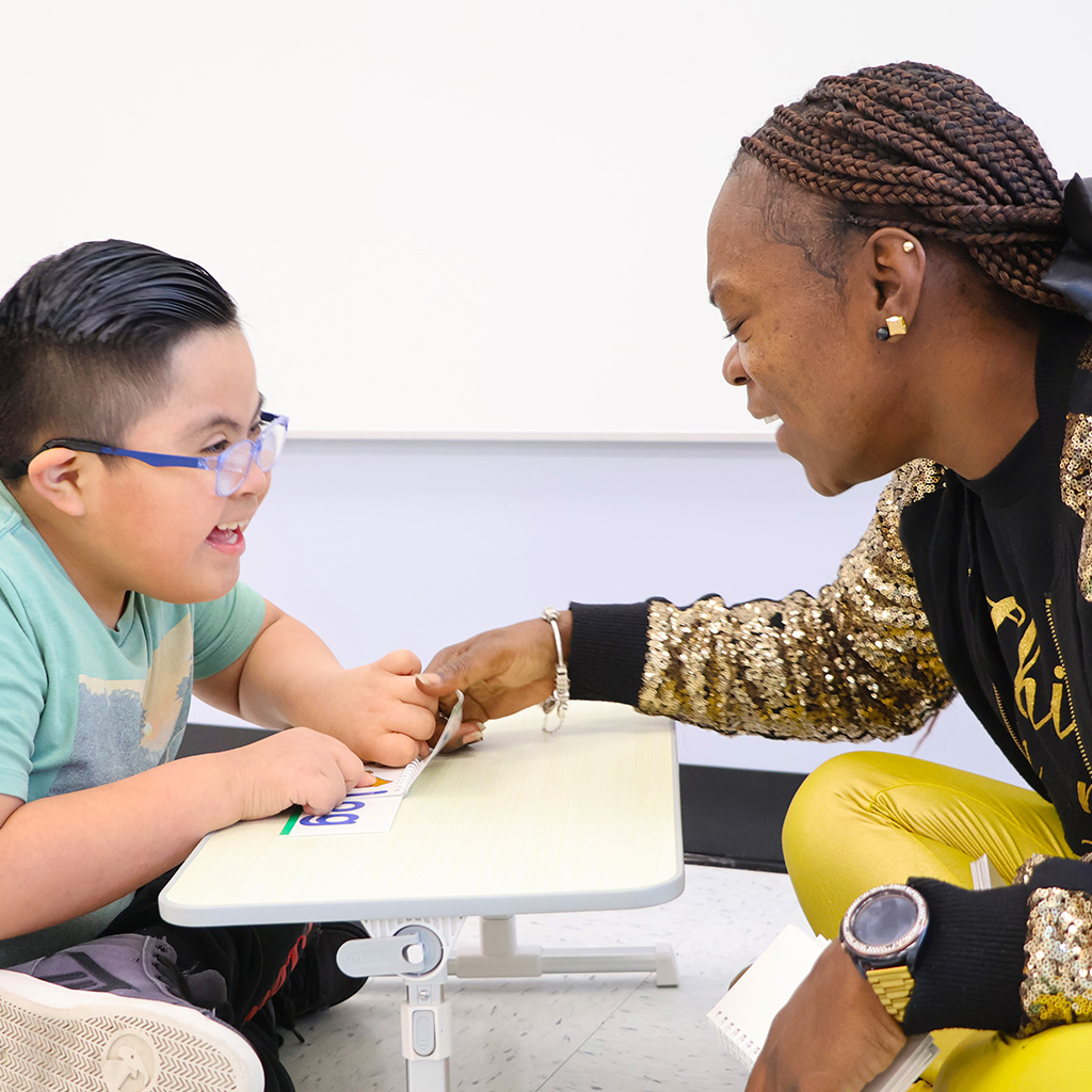 A boy and a woman sit at a desk practicing reading together.