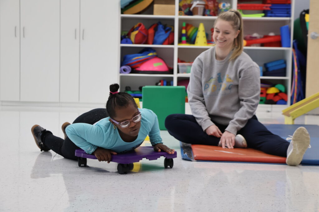 A girl on her stomach on a scooter with a physical therapist sitting on a mat next to her.