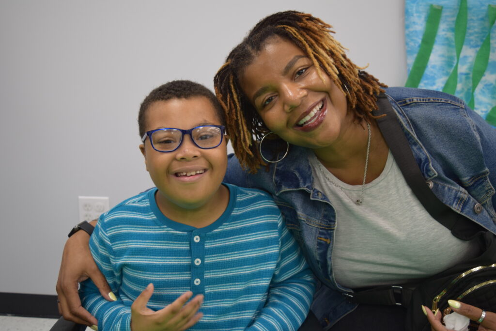 A boy and his mom sit side by side, with the mom hugging the boy's shoulder.