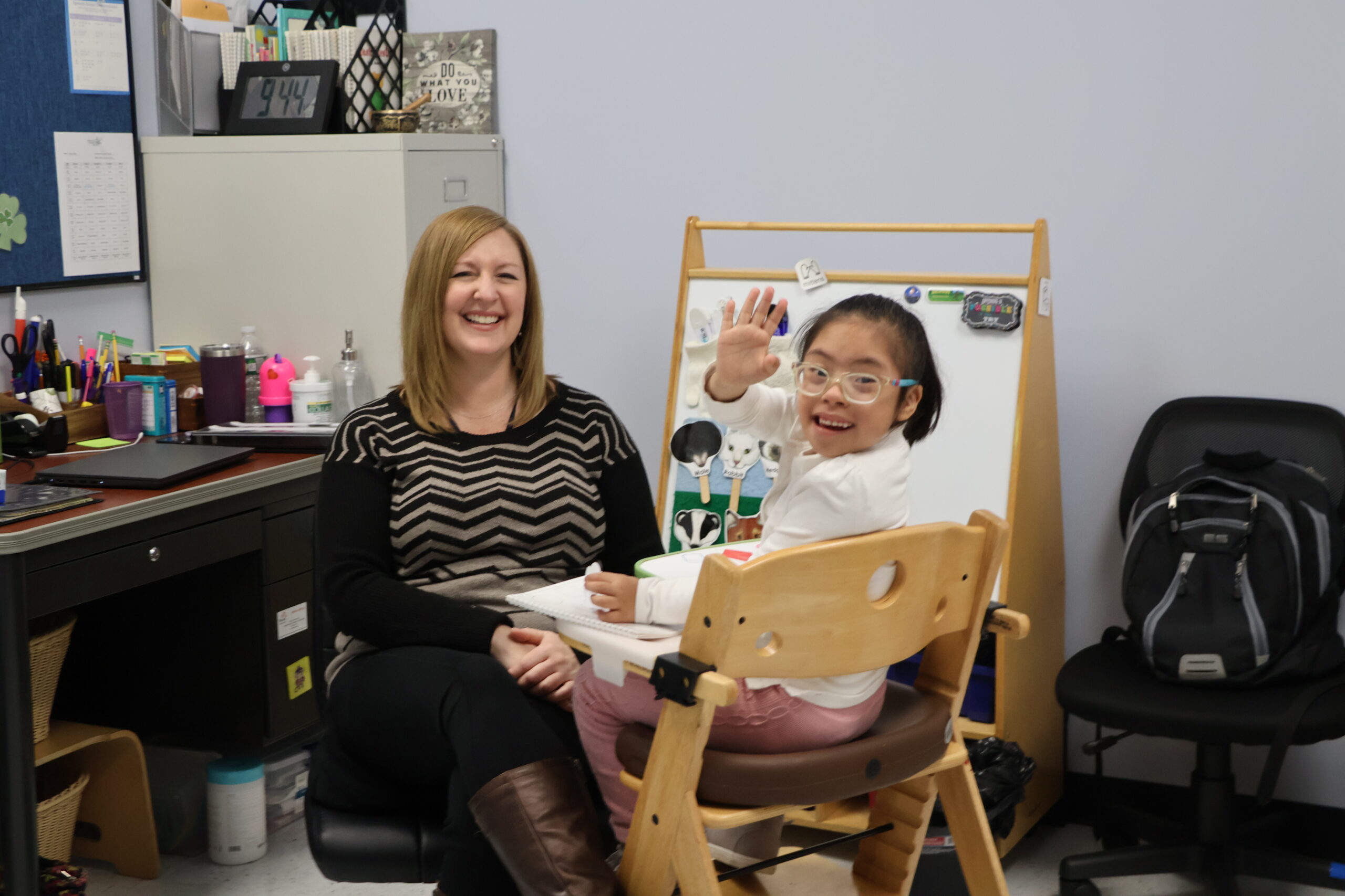 A speech therapist sits with a girl in glasses who is waving at the camera.